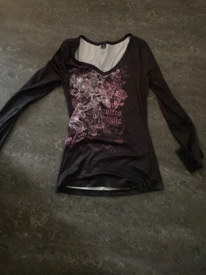 Svart långärmad topp med tryck - Svart långärmad topp med v-ringning och detaljerat rosa och vitt tryck framtill. Toppens material känns mjukt och stretchigt, perfekt för en cool och avslappnad stil. Snygg passform och unik design framtill.