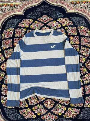 Randig långärmad topp Hollister - Säljer en långärmad topp från Hollister med breda blå och vita ränder. Toppen har rund halsringning och är tillverkad i mjuk bomull. Perfekt för en avslappnad och stilren look.