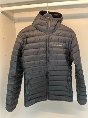 Svart dunjacka Peak Performance - Snygg svart dunjacka från Peak Performance med huva och dragkedja framtill. Jackan har två sidofickor med dragkedja och diskret logga på bröstet. Perfekt för kalla vinterdagar och riktigt lätt att bära.