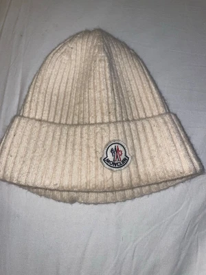 Vit ribbstickad mössa från Moncler - Snygg vit ribbstickad mössa från Moncler med uppvikt kant och klassisk Moncler-logga framtill. Mössan är tillverkad i mjukt material och har en stilren, enkel design som passar perfekt till kalla dagar.