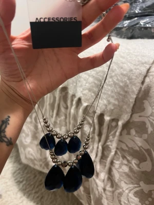 Silverfärgat halsband med blå droppar - Snyggt halsband från Accessories med silverfärgad kedja och stora blå droppformade hängen. Halsbandet har även silvriga pärlor som detaljer och ger en cool statement-look. Perfekt för att lyfta din outfit med lite extra färg och stil.