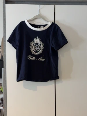 Mörkblå t-shirt Belle Âme H&M Divided - Mörkblå t-shirt från H&M Divided med vit krage och tryck framtill med texten 'Belle Âme' och ett dekorativt emblem. Kortärmad och i mjuk bomull, perfekt för en avslappnad och trendig look.