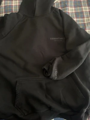 Svart Essentials Fear of God hoodie - Svart hoodie från Essentials Fear of God med diskret logga på bröstet och ärmen. Klassisk känguruficka framtill och stor huva. Mjuk och skön bomullsblandning, perfekt för en avslappnad streetwear-look.