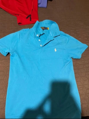 Blå pikétröja från Polo Ralph Lauren - Klassisk blå pikétröja från Polo Ralph Lauren med vit broderad logga på bröstet. Tröjan har korta ärmar, krage och knappar framtill. Perfekt till jeans eller shorts för en clean och stilren look. Storlek M men passar som S