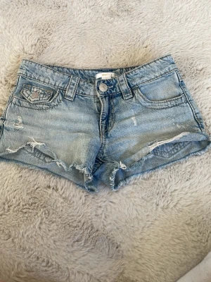 Blå jeansshorts med broderade fickor - Snygga ljusblå jeansshorts från gina trico med slitna detaljer och råa kanter. Baksidan har broderade fickor med paisleymönster och små nitar. Perfekta för sommaren och ger en cool, avslappnad vibe. I storlek 146. 