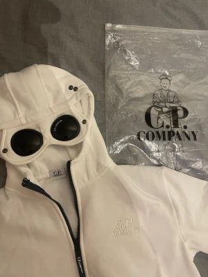 Vit hoodie med goggles från C.P. Company - Säljer en vit hoodie från C.P. Company med ikoniska inbyggda goggles i huvan och dragkedja framtill. Hoodien har broderad logga på bröstet och ribbade muddar. Materialet är mjukt och bekvämt, perfekt för en streetwear-look.