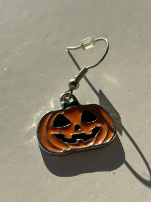 Halloween örhängen med pumpa - Coola örhängen med orange pumpaform och svart ansikte, perfekta för Halloween. Hängande modell i metall med färgglad emalj. Ger en lekfull och spooky vibe till din outfit.