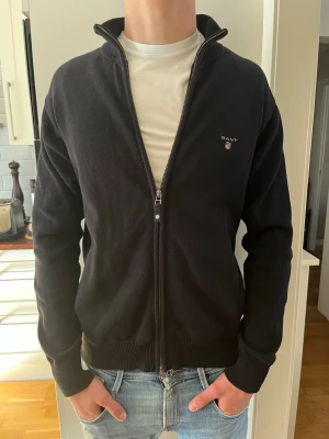 Svart ziptröja från Gant - Svart ziptröja från Gant med liten logga på bröstet. Tröjan har hög krage, ribbade muddar och nederkant samt dragkedja hela vägen. Perfekt till jeans och enkel att matcha. Materialet känns mjukt och bekvämt.