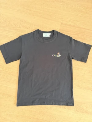 Svart t-shirt från Casablanca - Svart t-shirt från Casablanca med broderad logga och färgglad figur på bröstet. Klassisk rund halsringning och korta ärmar. Tillverkad i mjuk bomull som känns skön mot huden. Perfekt för en avslappnad och stilren look.