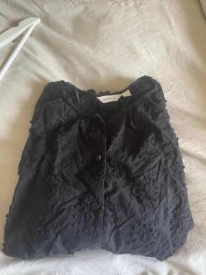Svart blommig blus från Dobber - Svart blus från Dobber med broderade blommor och knäppning framtill. Blusen har en lätt rynkad halsringning och är tillverkad i ett tunt tyg med struktur. Perfekt för dig som gillar detaljer och en stilren look. Långärmad 