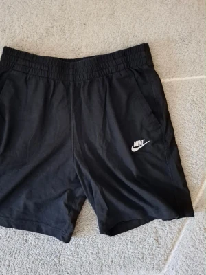 Svarta Nike shorts i bomull XL - Svarta shorts från Nike i storlek XL, tillverkade i mjuk bomull. Klassisk design med elastisk midja och sidofickor. Perfekta för träning eller chill dagar. Nike-logga i vitt på ena benet ger en sportig vibe.