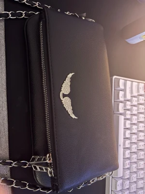 Zadig Voltaire väska - Snygg svart handväska i läderimitation med silverfärgad kedjerem och dragkedja. Väskan har en dekorativ detalj med silverfärgade vingar på framsidan och en tofs vid dragkedjan. Perfekt för att lyfta din outfit med en cool touch.