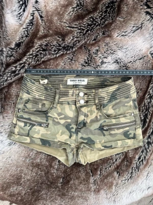 Camo shorts med dragkedjor Tally Weijl - Snygga camouflage-mönstrade shorts från Tally Weijl med hög midja och coola dragkedjedetaljer framtill. Shortsen har tre knappar i midjan och fickor med dragkedjor. Perfekta för en edgy och avslappnad stil.