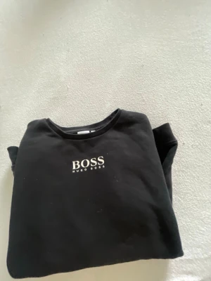 Svart sweatshirt från Hugo Boss - Svart sweatshirt från Hugo Boss med vit logga tryckt på bröstet och även på ryggen. Klassisk rund halsringning och långa ärmar. Perfekt för en stilren och avslappnad look.