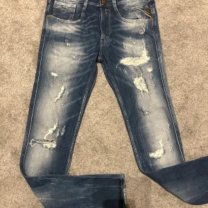 Replay jeans  - Säljer ett par Replay Anbass jeans i blå tvätt med slitna detaljer och snyggt distressed look. Klassisk femficksmodell med dragkedja och Replay-logga på fickan. Jeansen är i stretchig bomull och har en modern passform som sitter snyggt. (Dessa jeans är i jätte bra skicka men det är ett hål vid knät men är med design) skriv vid funderingar 