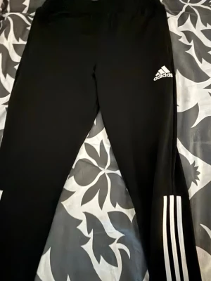 Svarta leggings från Adidas - Snygga svarta leggings från Adidas med klassiska vita ränder längs ena benet och Adidas-logga på låret. Tight passform och stretchigt material som passar perfekt till träning eller chill. Sportig look som alltid är trendig.