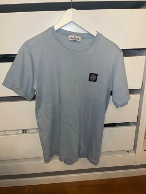 Ljusblå Stone Island t-shirt - Säljer en ljusblå t-shirt från Stone Island med klassisk logopatch på bröstet. T-shirten har rund hals och korta ärmar. Tillverkad i mjuk bomull för skön känsla och enkel stil. Ja den är äkta, köpt på nk för några år sendan. Storlek 170 men passar S