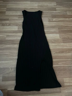 Svart ribbad långklänning med slits - Snygg svart långklänning i ribbat material med slits framtill. Klänningen är ärmlös och har en kroppsnära passform som framhäver figuren. Perfekt för dig som gillar stilrena och trendiga plagg. 