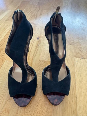 Svarta pumps från Enzo Angiolini strl 36 - Eleganta högklackade sandaler från Enzo Angiolini i svart mocka med dekorativa detaljer i sköldpaddsmönstrad finish och guldfärgade dragkedjor baktill. Strl 36, klackhöjd 9-10cm. I fint skick, används fåtal gånger 