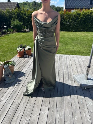 Balklänning  - Snygg grön axelbandslös långklänning med draperad överdel och broderade detaljer i midjan. Klänningen har en elegant omlottkänsla framtill och slits för extra rörelsefrihet. Perfekt för speciella tillfällen och ger en lyxig känsla. Klänningen är i en storlek 32 då den är insydd men dessa stygn är super lätta att ta bort vilket gör att den blir en 34. Den där från proms and weddings i Malmö och köptes för 2500 kronor plus hundra kronor frakt. Pris kan diskuteras!!!