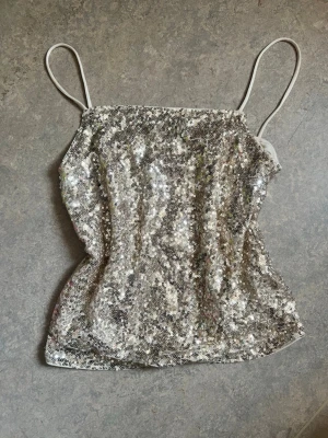 Silvrigt paljettlinne med smala band - Glittrigt linne i silver/guld med tunna axelband och täckt av paljetter. Perfekt för dig som vill sticka ut och älskar att glänsa. Rak passform och mjukt tyg på insidan för extra komfort.
