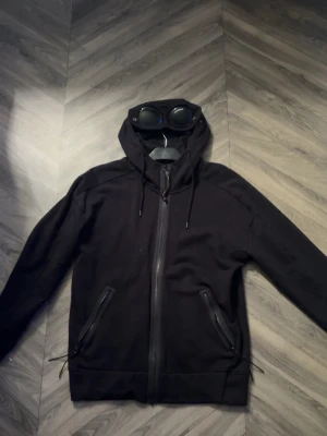 Cp company zip hoddie - Säljer en svart hoodie från C.P. Company med dragkedja framtill och två snedställda fickor med dragkedja. Huvan har inbyggda glasögon, vilket ger en unik och streetinspirerad look. Tillverkad i mjukt bomullsmaterial och har justerbara snören i huvan. Kan fixa fler bilder privat tveka inte på o höra av dig vid frågor