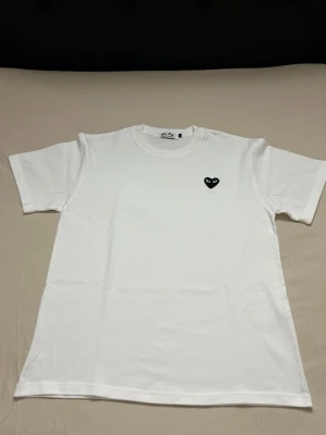 Vit Comme des Garçons Play t-shirt - Säljer en klassisk vit t-shirt från Comme des Garçons Play med det ikoniska svarta hjärtat med ögon broderat på bröstet. T-shirten har rund halsringning och är tillverkad i mjuk bomull. Perfekt basic-plagg med en stilren twist.