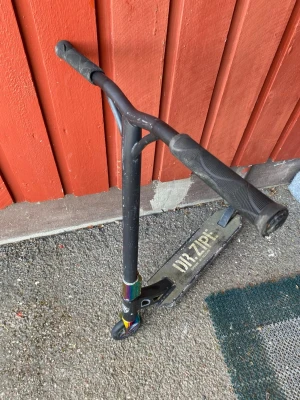 Svart trick sparkcykel Dr. Zipe - Säljer en svart trick sparkcykel från Dr. Zipe med robust metallram, breda handtag och regnbågsfärgad clamp och framgaffel. Perfekt för skateparken eller att cruisa runt på. Deck i metall med Dr. Zipe-logga och slitstarka hjul.
