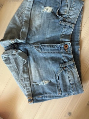 Blå denimshorts med slitningar - Säljer ett par blå denimshorts med coola slitningar och klassiska femficksdetaljer. Shortsens ben är uppvikta och de har en knappstängning framtill. Perfekta för dig som gillar en avslappnad och trendig look.