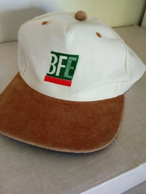 Vit och brun keps med BFE-brodyr - Säljer en unik keps med vit bomullstopp och brun sammetsliknande skärm. Framtill finns en brodyr med texten 'BFE' i grönt och vitt, samt en röd linje under. Klassisk form med knapp på toppen och justerbar passform.