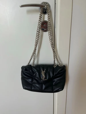 Svart YSL quiltad handväska - Säljer en svart quiltad handväska från Saint Laurent med silverfärgad kedjerem och ikonisk YSL-logga framtill.                                                                                 !!Jätteviktigt den är inte  @kta!!