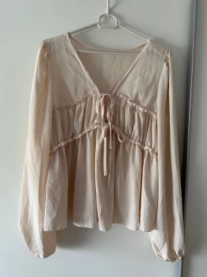 Ljusbeige blus med knytband - Supersöt ljusbeige blus med v-ringning och knytband framtill. Blusen har volangdetaljer över bysten och vida, långa ärmar med resår vid ärmslut. Tillverkad i ett tunt, flowy material som ger en romantisk vibe.