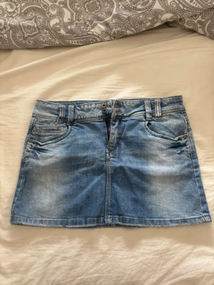 Ljusblå jeanskjol med broderade fickor - Säljer en ljusblå jeanskjol i kort modell med klassiska fem fickor och bälteshällor. Kjolen har snygga broderade detaljer på bakfickorna och stängs med dragkedja och dubbla knappar framtill. Perfekt till sommaren och enkel att matcha.