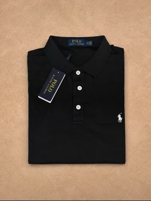 Svart pikétröja från Ralph Lauren - Klassisk svart pikétröja från Ralph Lauren med vit broderad logga på bröstet. Tröjan har korta ärmar, krage och tre vita knappar framtill. Tillverkad i mjuk bomull som känns skön mot huden. Perfekt till jeans eller shorts för en clean look.