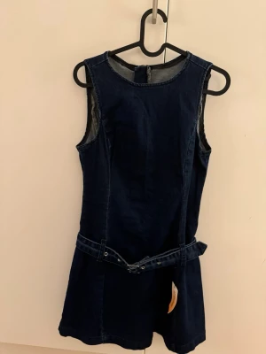 Mörkblå ärmlös jeansklänning med bälte - Mörkblå klänning i jeansmaterial med ärmlös design och rund hals. Klänningen har ett matchande bälte i midjan som ger extra stilpoäng. Enkel och trendig modell som är lätt att styla till olika looks.