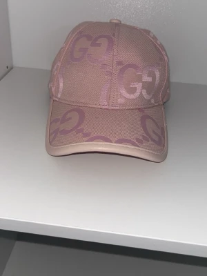 Rosa keps från GG med GG-mönster - Säljer en snygg rosa keps från GG med det klassiska GG-mönstret i ljusrosa och lila toner. Kepsen har böjd skärm och är tillverkad i ett tåligt tygmaterial med detaljer i konstläder runt skärmen. Perfekt för dig som gillar exklusiva accessoarer.