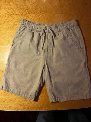 Beige shorts med snörning - Säljer ett par beige shorts med avslappnad passform och snörning i midjan. Shortsen har två sidofickor och är tillverkade i ett mjukt material som känns skönt mot huden. Perfekta för varma dagar och enkel att matcha med olika stilar. Från Kappahl i storlek 158.