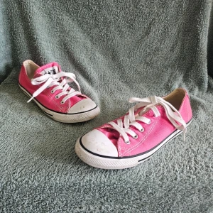 Rosa Converse All Star låga sneakers - Klassiska Converse All Star sneakers i starkt rosa canvas med vita gummisulor och tåhätta. Skorna har vita snören och svarta detaljer längs sulan. Perfekta för en avslappnad och cool stil. Ikonisk logga på plösen och tidlös design.