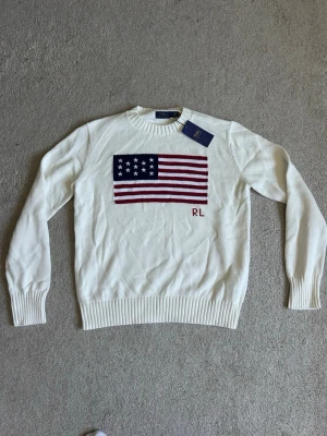 Vit stickad tröja Polo Ralph Lauren - Vit stickad tröja från Polo Ralph Lauren med amerikansk flagga i rött, vitt och blått på bröstet samt RL-broderi. Tröjan har rund halsringning och ribbstickade muddar vid ärmslut och nederkant. Perfekt för dig som gillar klassisk preppy stil.