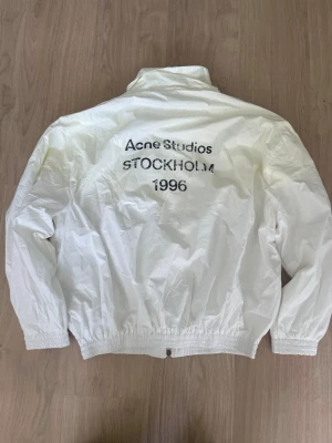 Vit vindjacka Acne Studios - Snygg vit vindjacka från Acne Studios med tryck på ryggen där det står 'Acne Studios STOCKHOLM 1996'. Jackan har dragkedja framtill, hög krage och resår vid ärmslut och midja. Perfekt för en clean och stilren look.