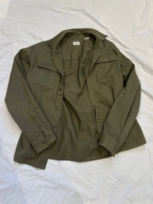 Olivgrön overshirt från Jack & Jones  - Stilren olivgrön overshirt från Jack& Jones  med två bröstfickor och klassisk krage. Tillverkad i ett mjukt bomullstyg och har knäppning framtill samt vid ärmsluten. Perfekt lager-på-lager-plagg för en avslappnad look.