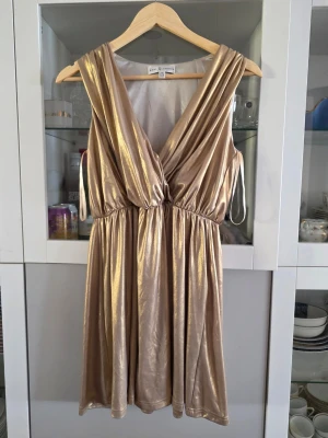 Guldig omlottklänning Rare London - Snygg guldig omlottklänning från Rare London med djup v-ringning och glansigt material. Klänningen har resår i midjan och är ärmlös med ett elegant fall. Perfekt för dig som vill sticka ut med metallic-look. Köpt inför temafest men användes aldrig, ingen lapp kvar men ny