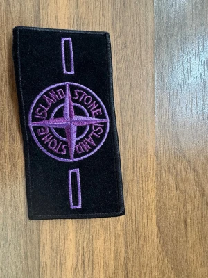 Stone island badge purple - Säljer en svart patch från Stone Island med broderad kompasslogga i lila. Märket är rektangulärt och har två öppningar för fastsättning. Perfekt för att sätta på jacka, tröja eller väska för en unik look.