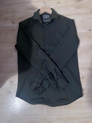 Mörkgrön skjorta från Fusaro - Stilren mörkgrön skjorta från Fusaro med klassisk krage och knappar framtill. Skjortan har lång ärm och är tillverkad i ett mjukt material som passar perfekt till olika outfits. Enkel och clean design utan mönster.