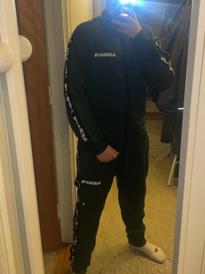 Grön svart vit dress från askari  - Mysigt svart loungewear-set från Asika med vita logotyper på bröstet och benet samt text längs ärmar och byxben. Setet består av en zip-jacka och matchande byxor i mjukt syntetmaterial, perfekt för chill hemma.