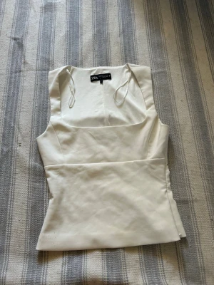 Vit ärmlös topp från Zara - Snygg vit topp från Zara med fyrkantig ringning och smala axelband. Toppen är ärmlös och har en figurnära passform med dold dragkedja i sidan för enkel påklädning