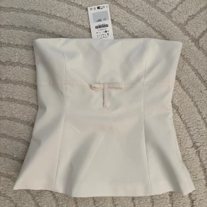 Axelbandslös topp med rosett - Vit axelbandslös topp med rosett, lite peplum liknande modell. helt ny med prislapp, Endast provad. Säljer pga att den inte passade mig🩷 