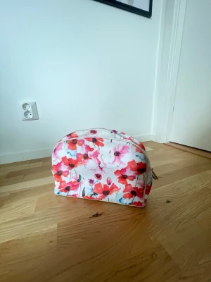 Blommig necessär med dragkedja - Söt necessär med blommigt mönster i rosa, rött och vitt från Lindex. Väskan har rundad form och dragkedja upptill. Insidan är ljusrosa med små prickar och flera fack för organisering. Perfekt för smink eller småprylar. Använd 