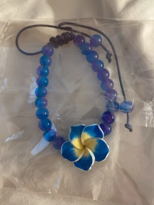 Blått pärlarmband med blomma - Armband med blå och lila pärlor samt en stor blå och gul blomma i mitten. Justerbart band i mörkblått. Perfekt för dig som gillar färgglada accessoarer och vill sticka ut med en tropisk vibe.