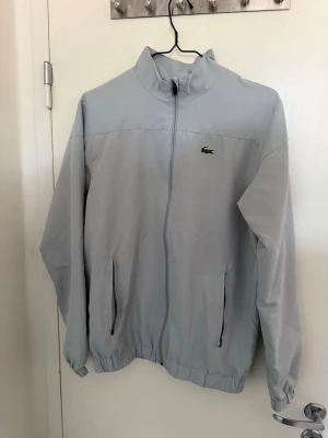 Ljusblå vindjacka från Lacoste - Snygg ljusblå vindjacka från Lacoste med dragkedja framtill, två sidofickor med dragkedja och klassisk ståkrage. Jackan har det ikoniska gröna krokodilmärket på bröstet och elastiska muddar vid ärmar och nederkant. Perfekt för blåsiga dagar.
